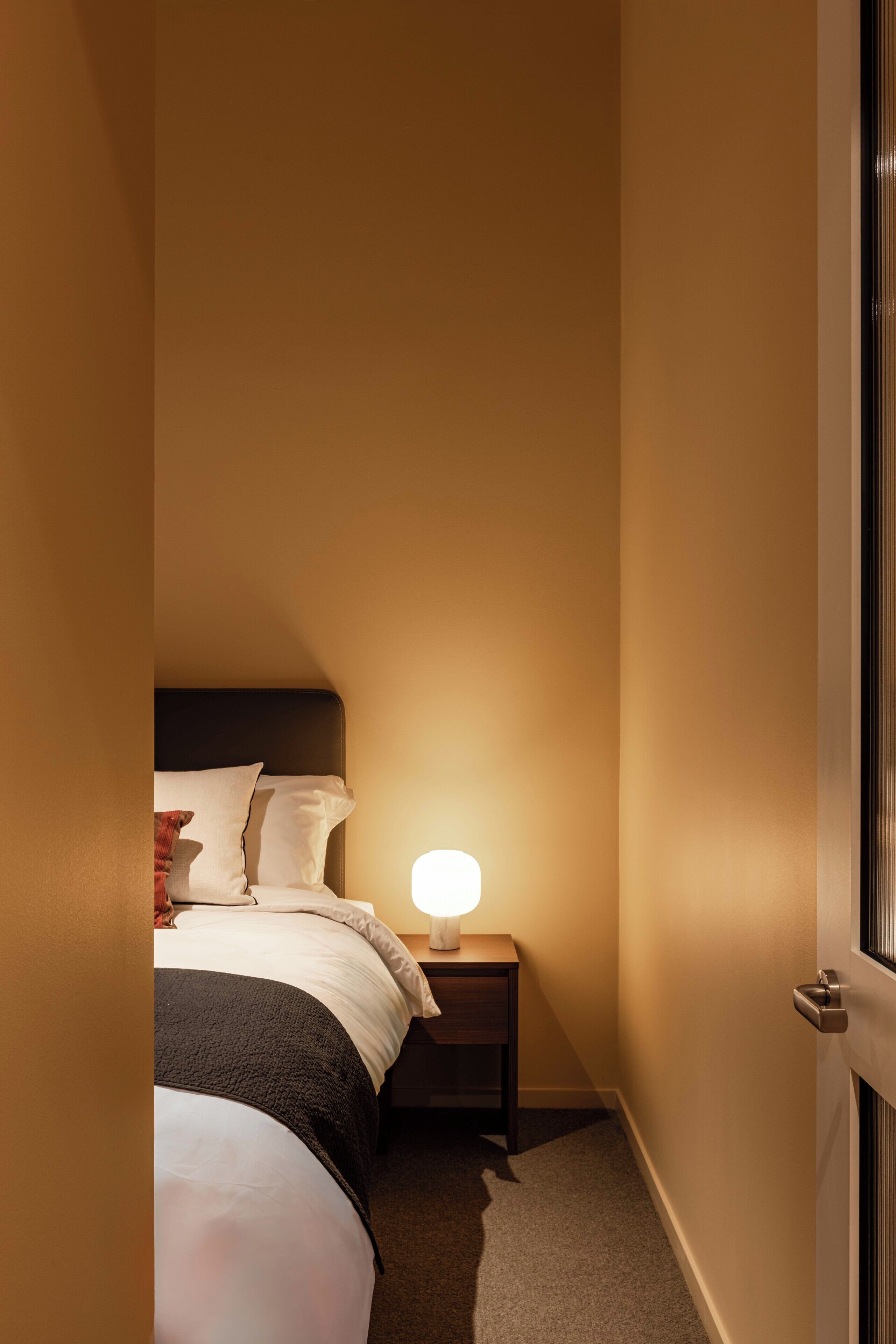 A narrow hotel room with a bed, bedside table, and a nightstand with a lamp.