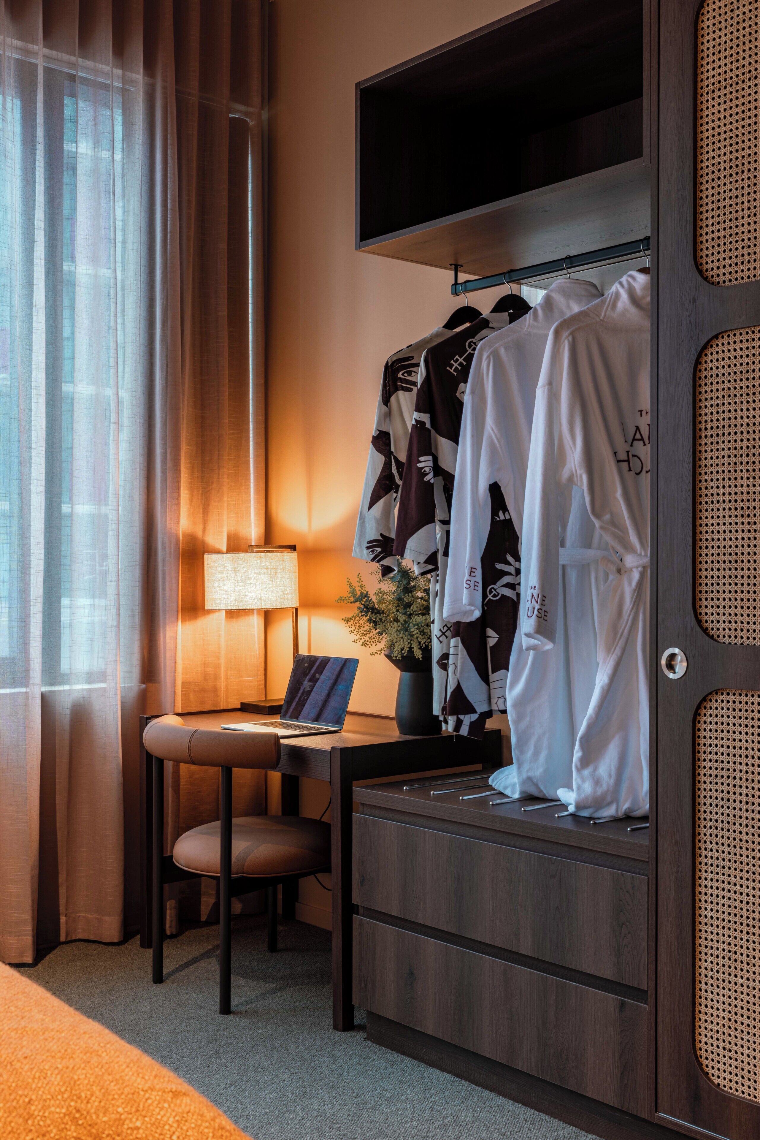 Ett hotellrum med en säng, ett skrivbord med en bärbar dator, en stol, en tv som är monterad ovanför en garderob och ett fönster med gardiner.