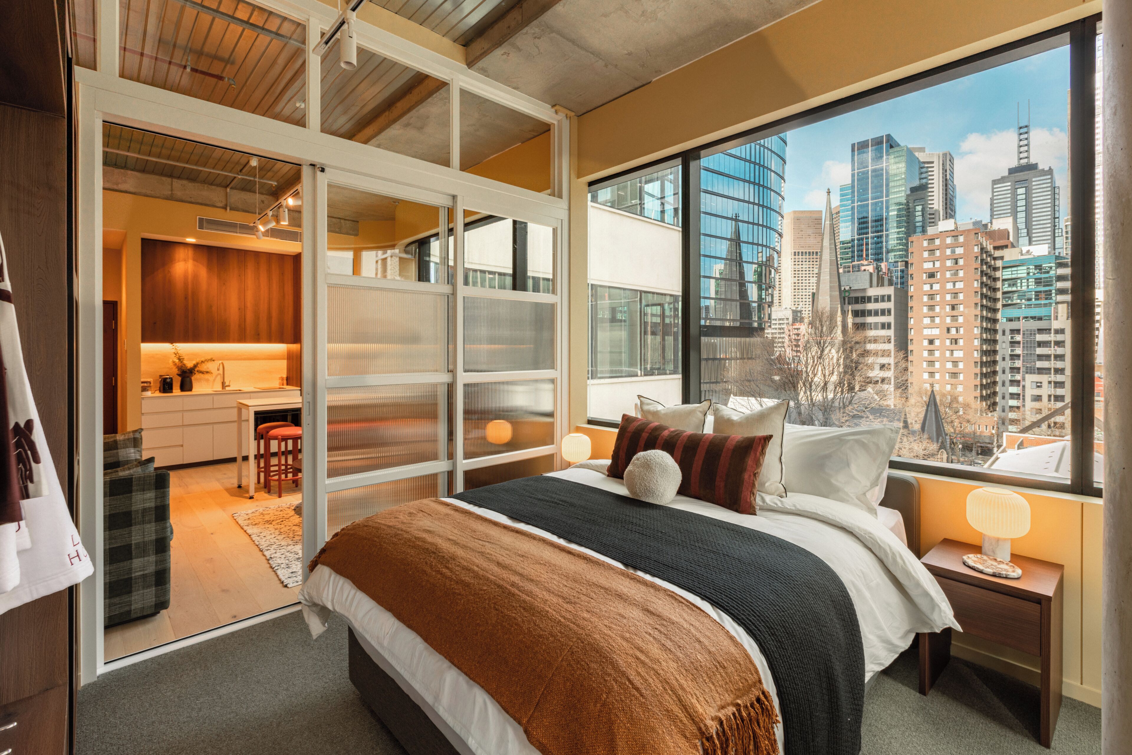 A modern bedroom with a large bed, a city view, and a kitchenette in the adjacent space.