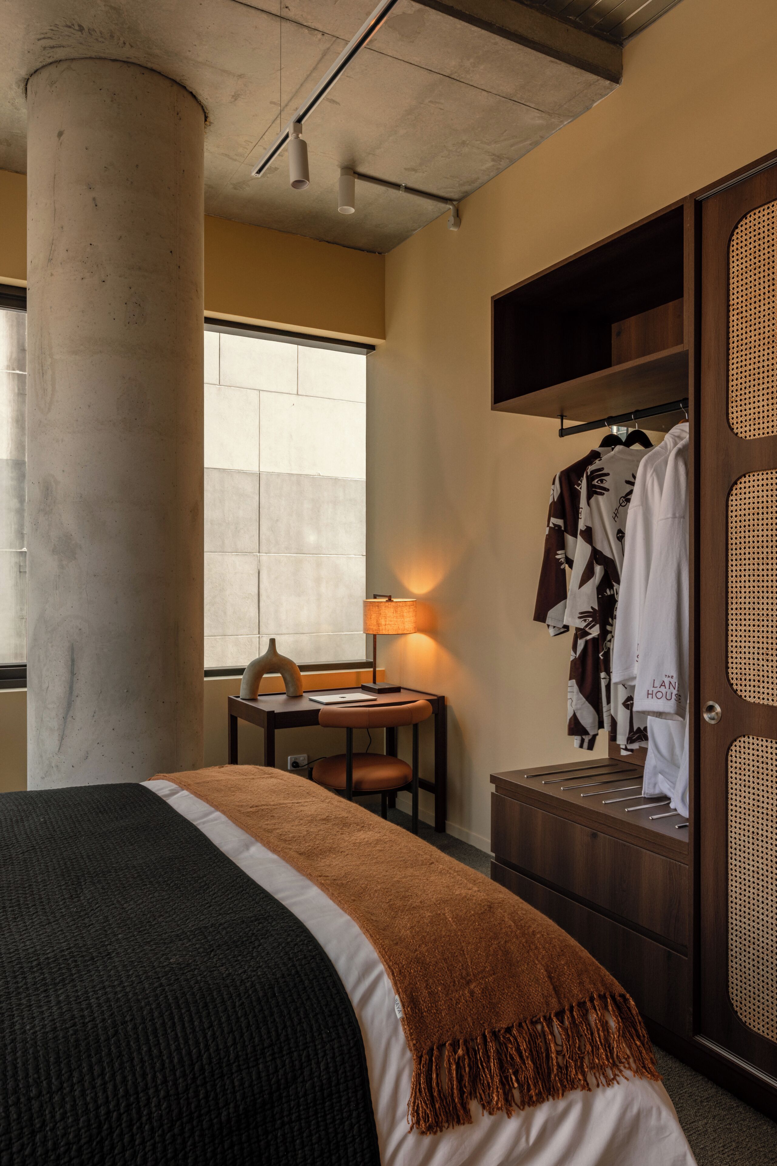 A hotel room with a bed, a desk with a lamp, a wardrobe, and a window.