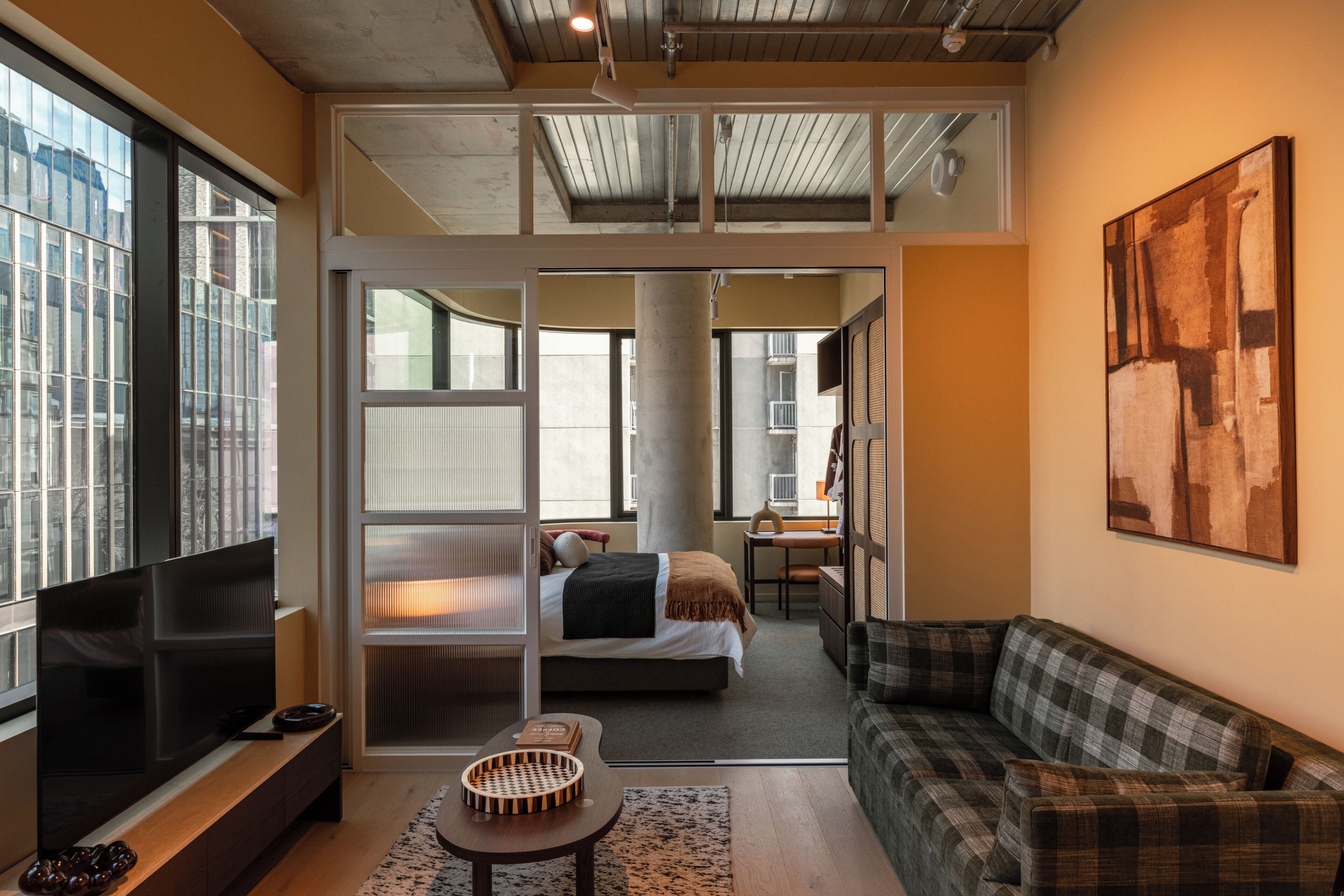 A modern living room with a flat-screen TV, a sofa, a dining area, and a bedroom with a bed and a desk.