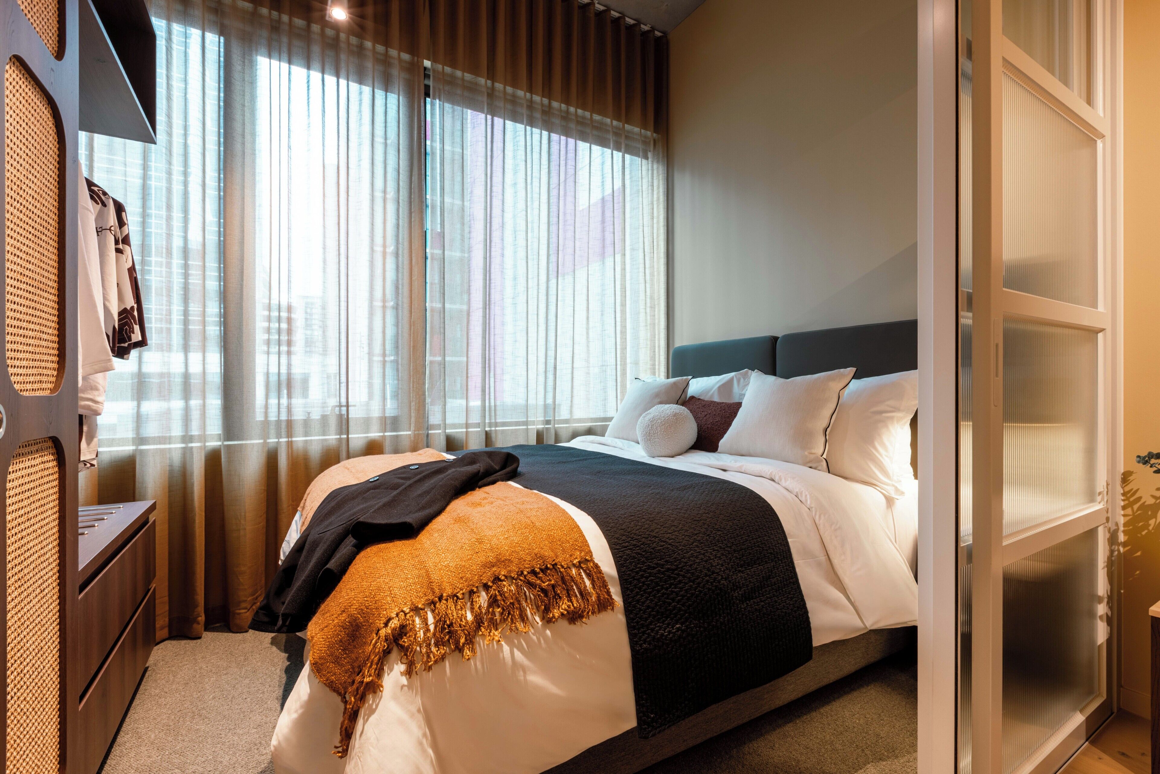 A hotel room with a bed, a wardrobe, a window with curtains, and a view of the city.