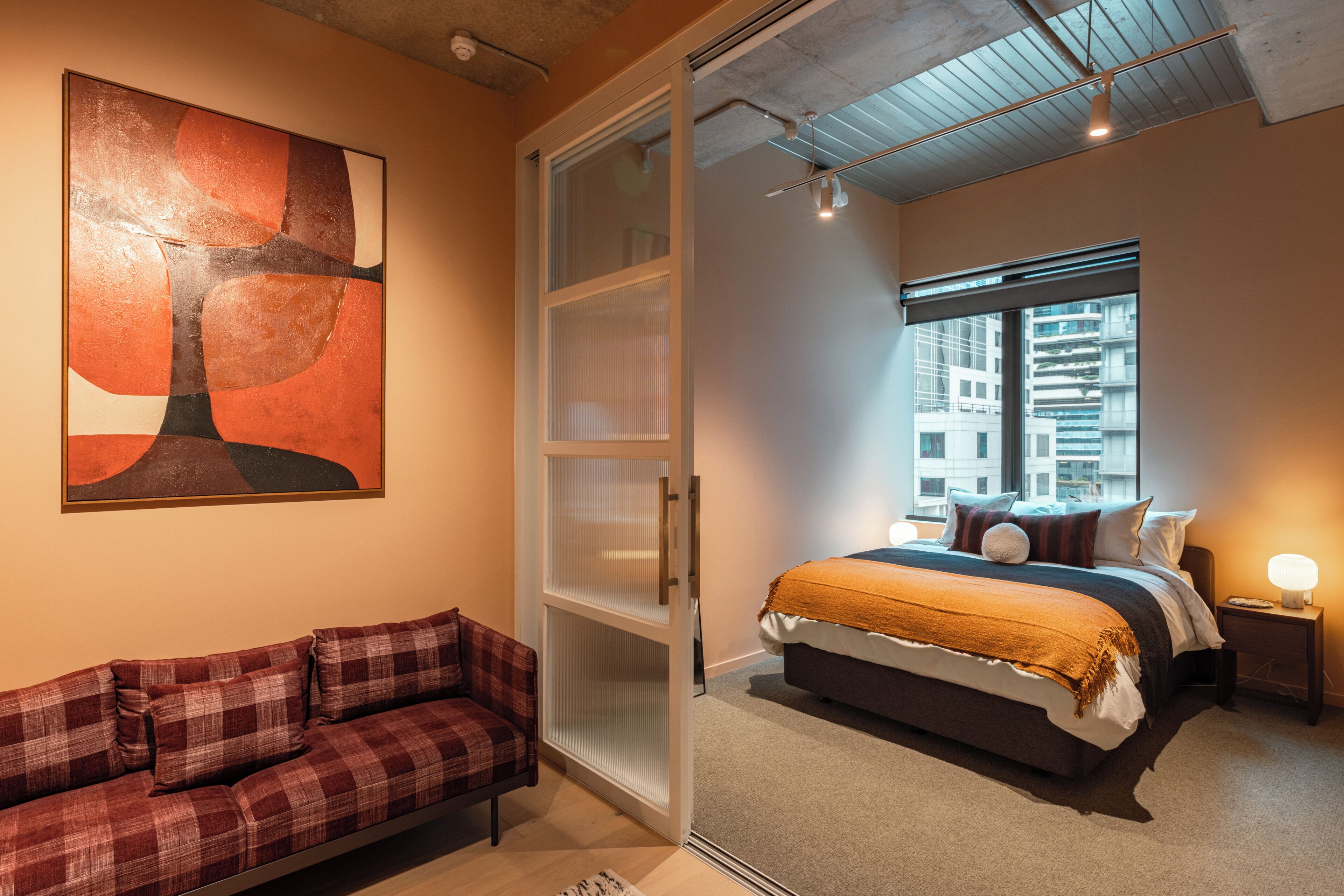A bedroom with a bed, a nightstand, a lamp, a window with a view of buildings, a sofa, and a painting on the wall.