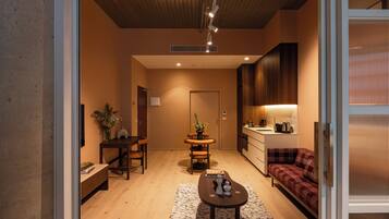 A modern living room with a wooden floor, a sofa, a dining table with chairs, and a kitchen area with cabinets and a sink.