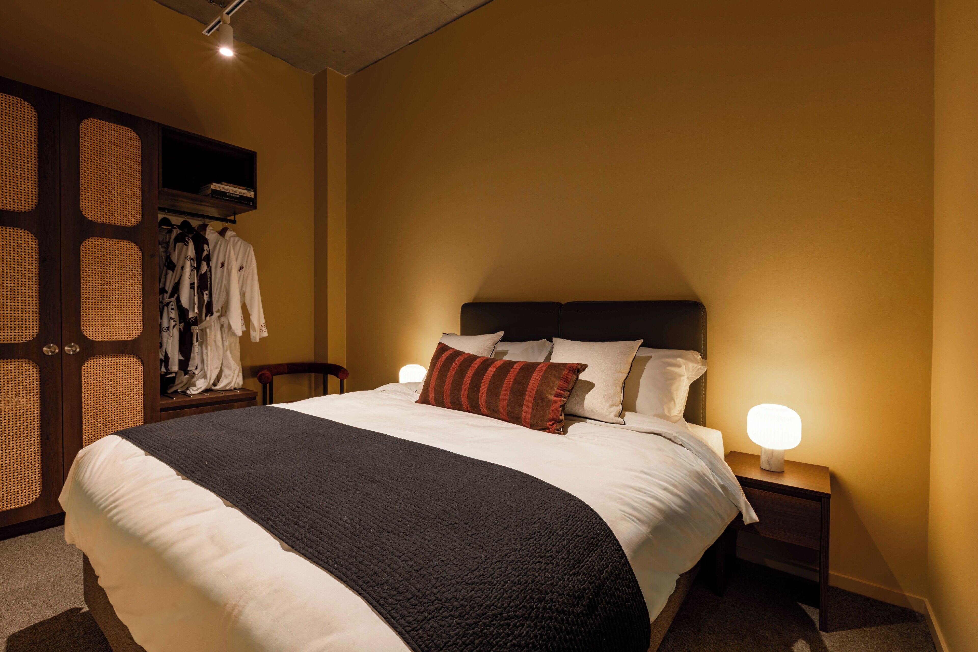 A neatly made bed with a black and white bedspread, two bedside lamps, and a wooden wardrobe with a woven design.