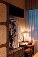 A hotel room with a wooden desk, a laptop, a lamp, and a clothing rack with robes.