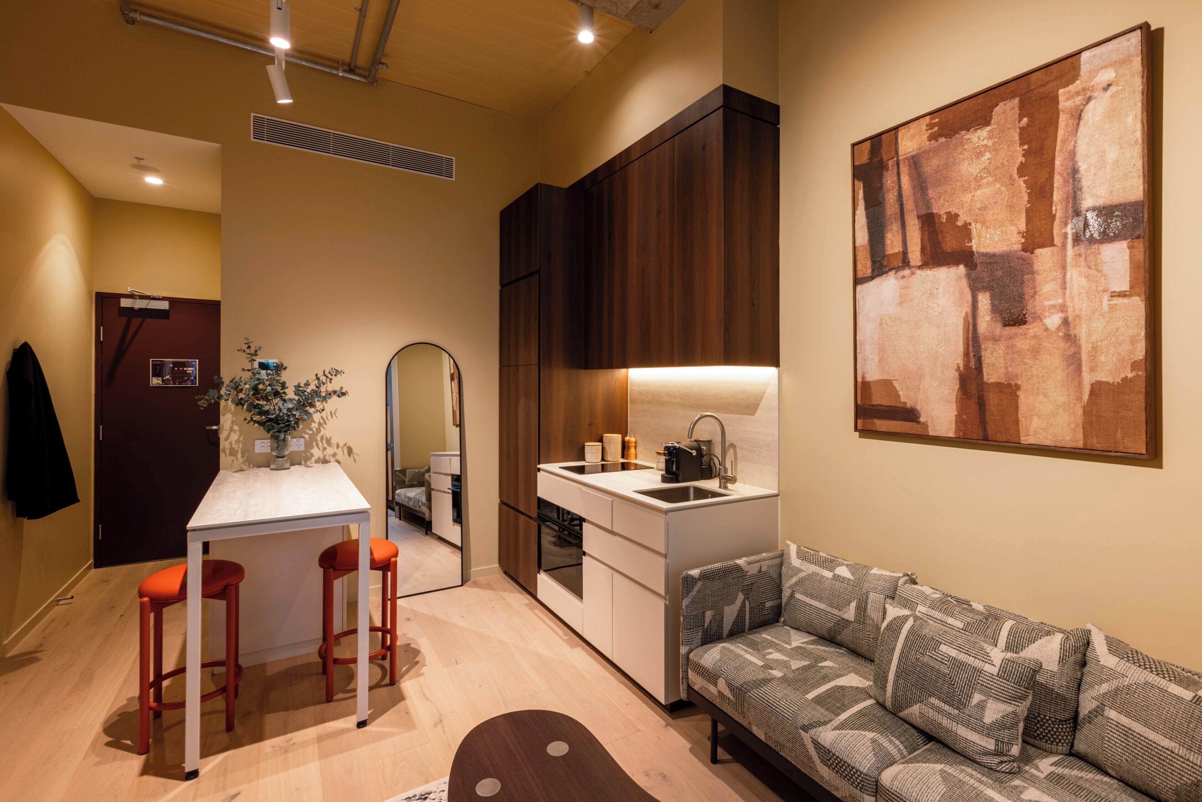A compact living space with a kitchenette, a dining area with stools, a sofa, and a large abstract painting on the wall.
