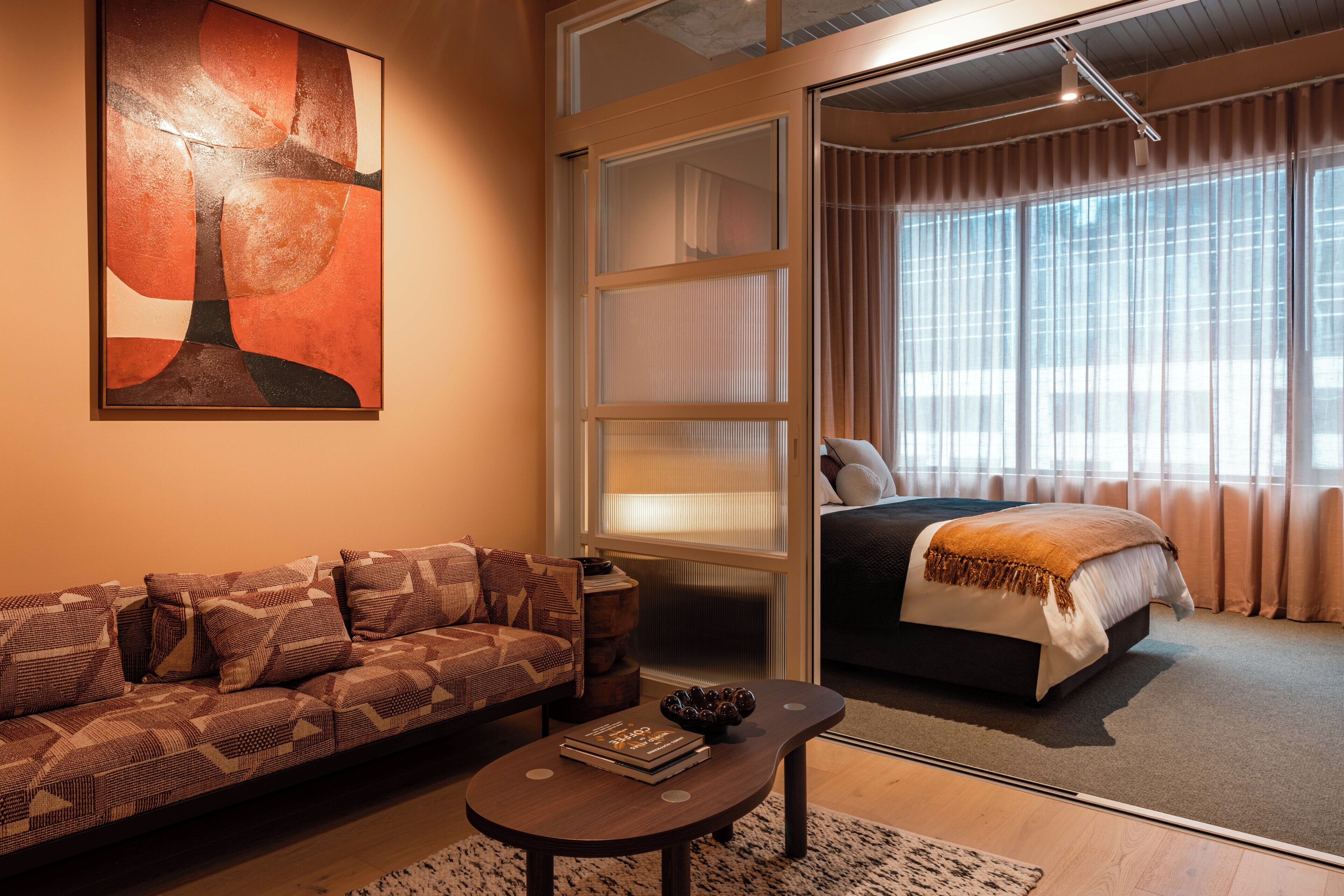 Habitación de hotel moderna con sofá, una mesa de centro, un cuadro abstracto grande, cama con vistas y una puerta corredera.