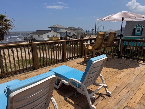 A wooden deck with lounge chairs and an umbrella, overlooking a beach with houses in the background.