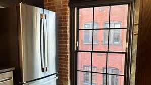 Una cocina con un refrigerador de acero inoxidable, una ventana con vista a un edificio de ladrillo y una mesa de madera.