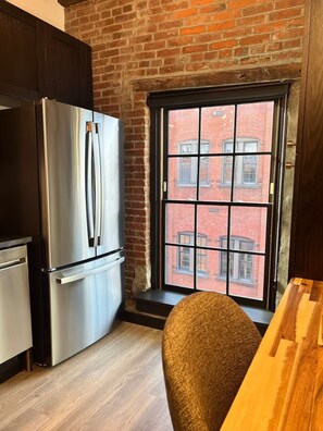 A kitchen with a stainless steel refrigerator, a window with a view of a brick building, and a wooden table. - Brick & Bed - Historic Loft/Walk to Stadium/Arch (St. Louis)