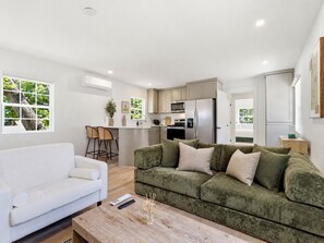 Ein modernes Wohnzimmer mit einem weißen Sofa, einem grünen Sofa mit Kissen, einem Couchtisch aus Holz und einer Küchenzeile mit Kühlschrank und Mikrowelle.