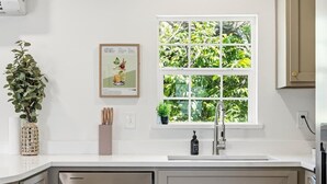 Eine Küche mit weißer Arbeitsplatte, einem Fenster mit Blick ins Grüne, einer gerahmten Speisekarte und einem Blumentopf.