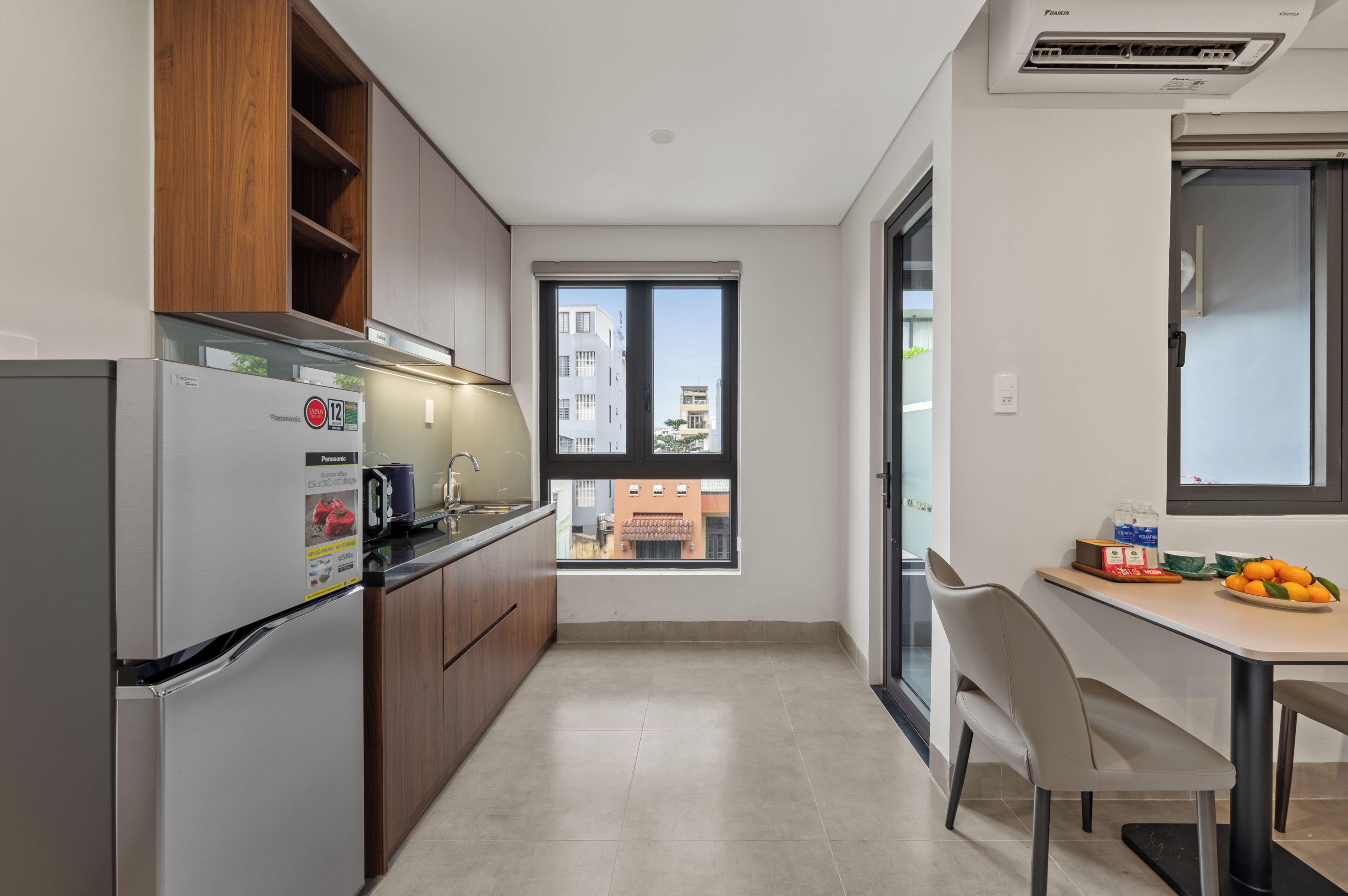 A modern kitchen with a refrigerator, wooden cabinets, a window with a view of buildings, and a dining area with a table and chairs.