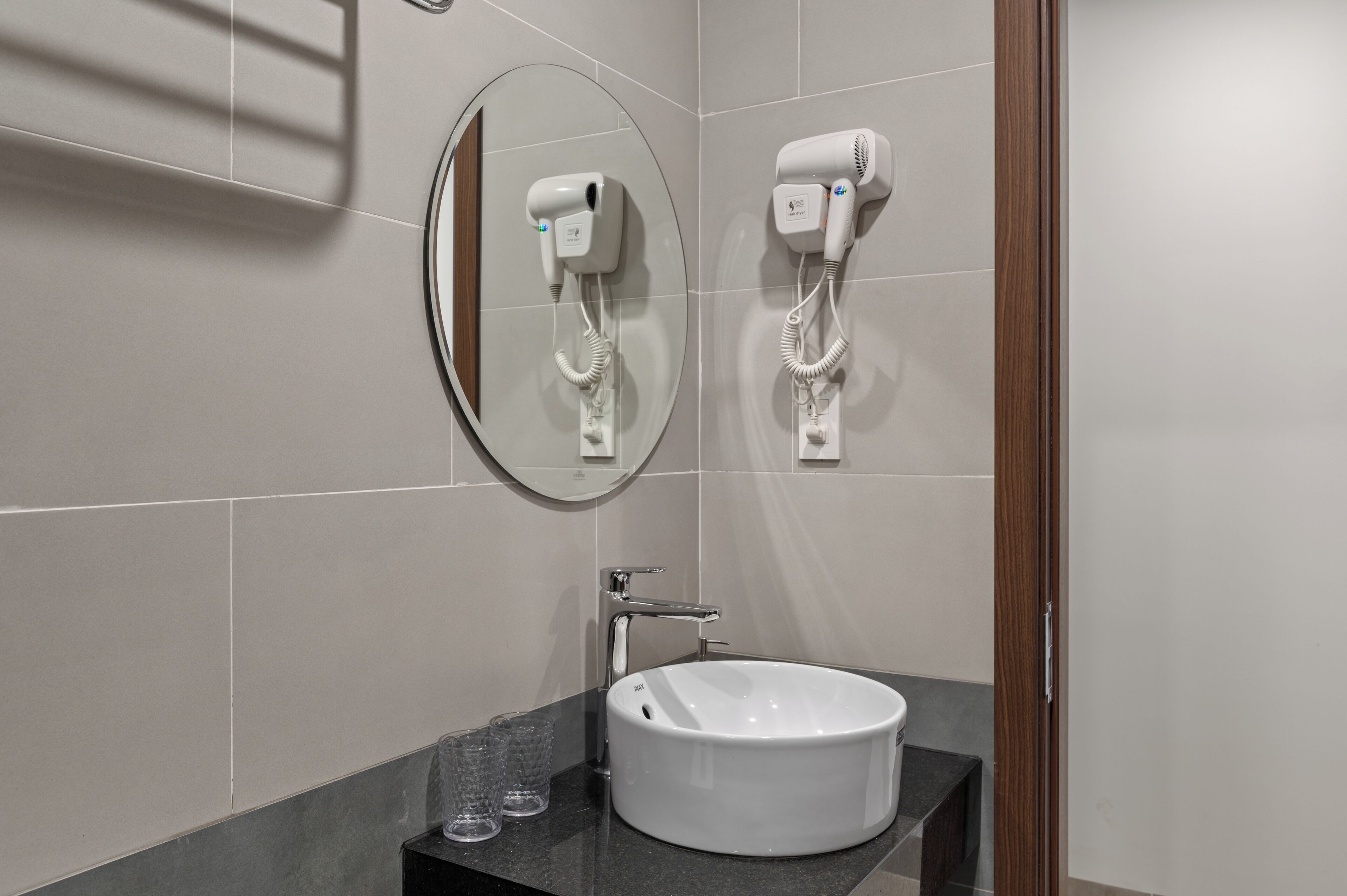 A bathroom with a round mirror, two hair dryers mounted on the wall, a white sink, and a dark countertop.