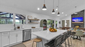 A modern kitchen with white cabinetry, a central island, and a dining area with a TV displaying a Disney castle. - Waterfront Paradise | Sauna, Hot Tub, Fire Pit, Private Dock & Water Crafts (Pocono Summit)