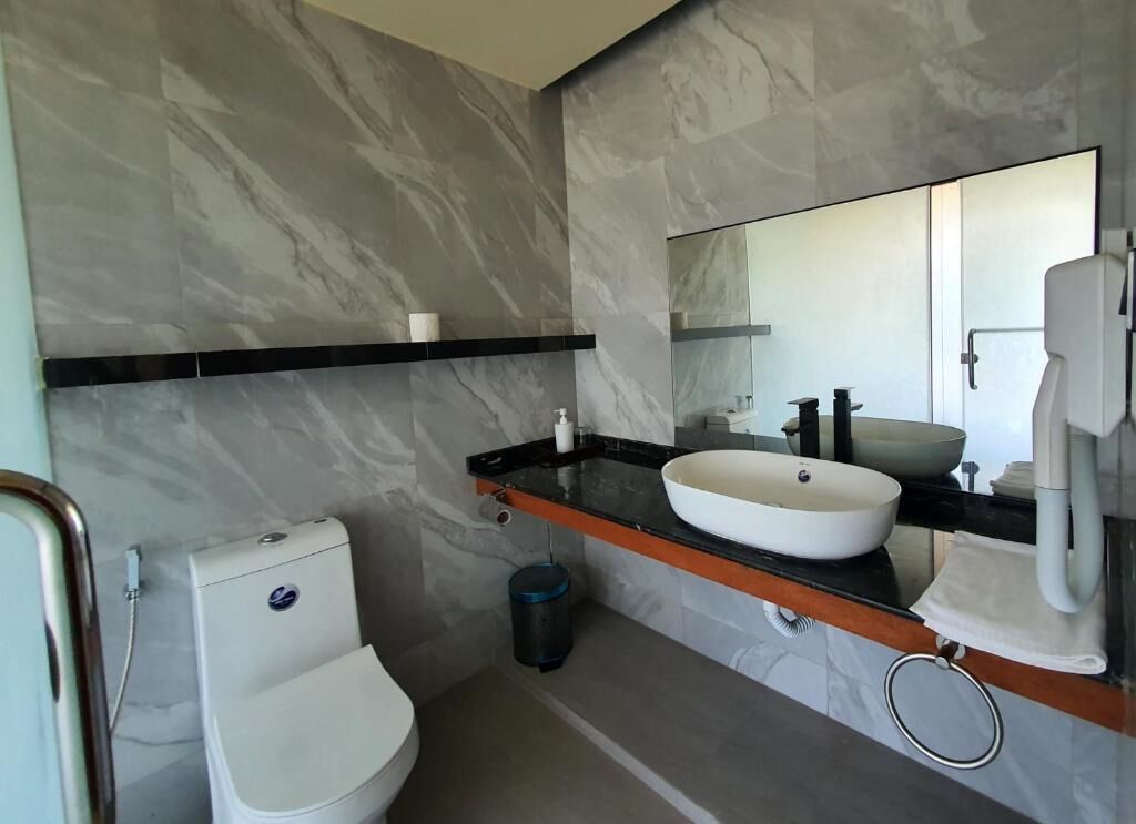Un bany modern amb una paret de marbre, un vàter blanc, una superfície de lavabo negra i un rentapeus blanc, juntament amb un mirall.