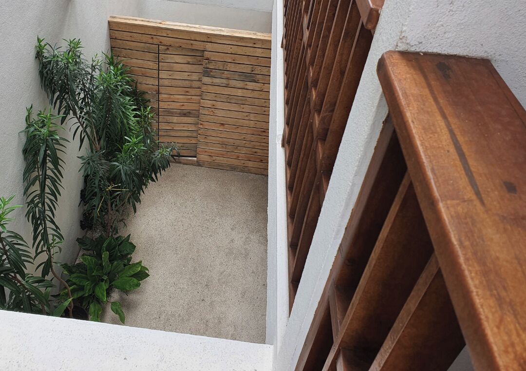 Un espacio exterior estrecho con cerca de madera, plantas en macetas y piso de concreto.