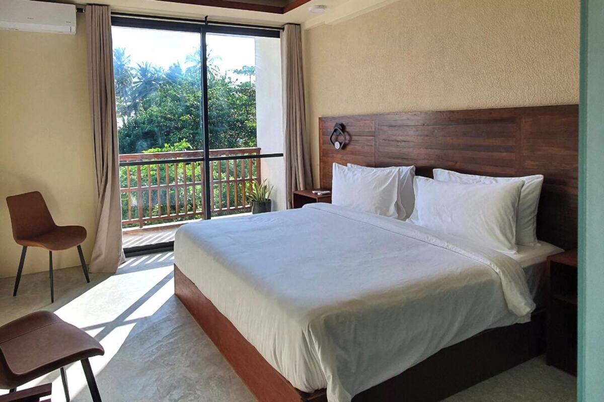 A hotel room with a large bed, a chair, a nightstand, and a balcony with a view of trees.