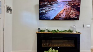 A living room with a flat-screen TV mounted on the wall, a fireplace below, and a small table with a plant in front.