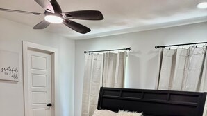 A bedroom with a black bed frame, a ceiling fan, a white door, and light-colored curtains.