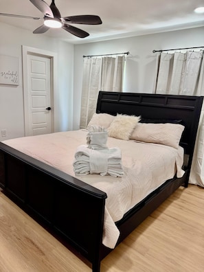A bedroom with a black bed frame, a ceiling fan, a white door, and light-colored curtains.