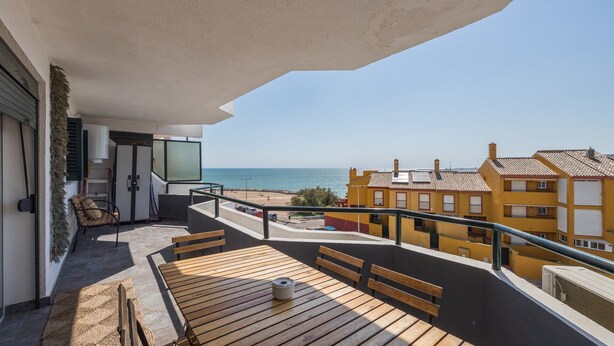 Un balcón con una mesa y bancos de madera, con vista a una playa y edificios amarillos.