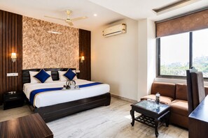 Quarto de hotel com cama, escrivaninha, cadeira, ventilador de teto, ar condicionado e janela com vista para a cidade.