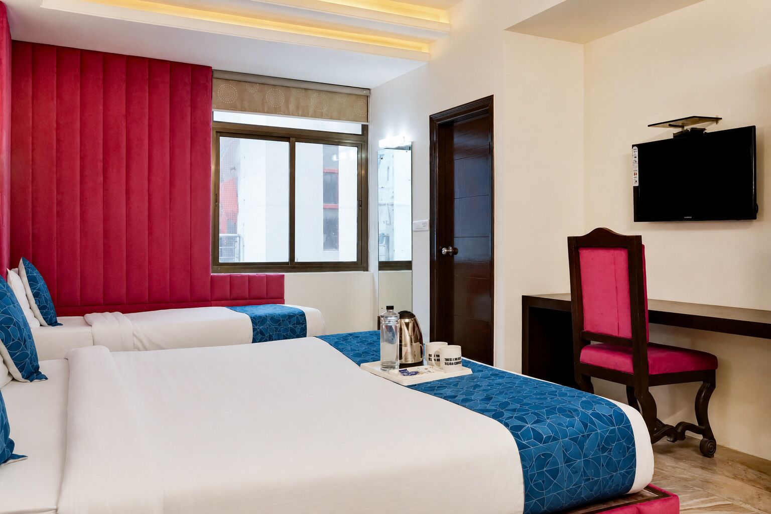 A hotel room with two beds, a desk, a chair, a TV, and a window with a view of buildings.