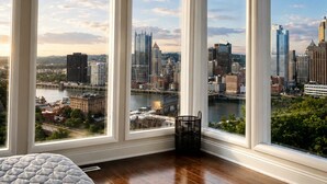A bedroom with a large bed, a view of a cityscape through large windows, and a wooden floor.