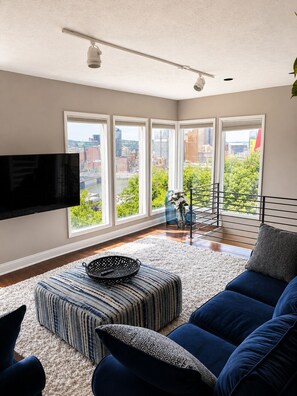 A modern living room with a large TV, a sofa, a patterned ottoman, and a view of the city through large windows.