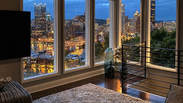 A room with a large window overlooking a cityscape at dusk, a flat-screen TV, a patterned rug, and a staircase with a railing.
