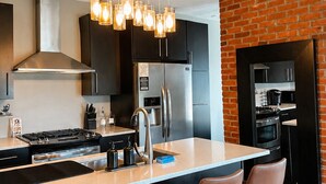 A modern kitchen with a central island, bar stools, stainless steel appliances, and a brick accent wall.