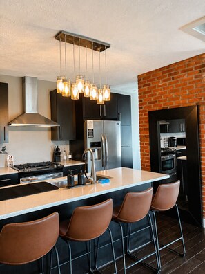 A modern kitchen with a central island, bar stools, stainless steel appliances, and a brick accent wall.