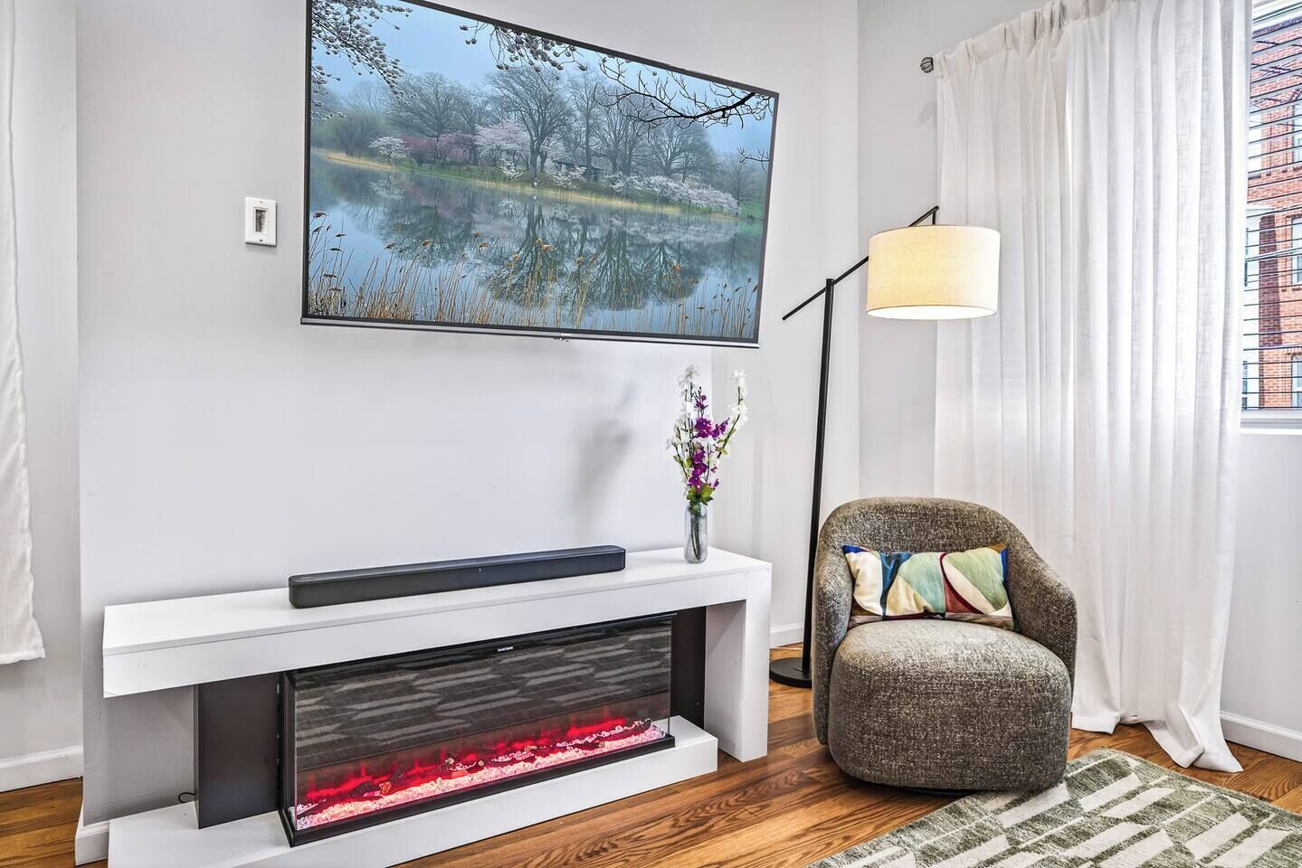 Een woonkamer met een open haard, een televisie erboven, een fauteuil met kussens en een vloerlamp.