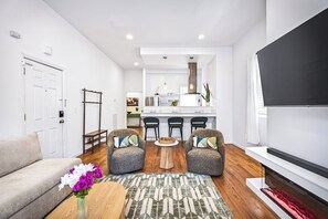 Ein modernes Wohnzimmer mit einer Couch, zwei Sesseln, einem Couchtisch und einem Fernseher. Im Hintergrund ist eine Küche mit einer Bar erkennbar.