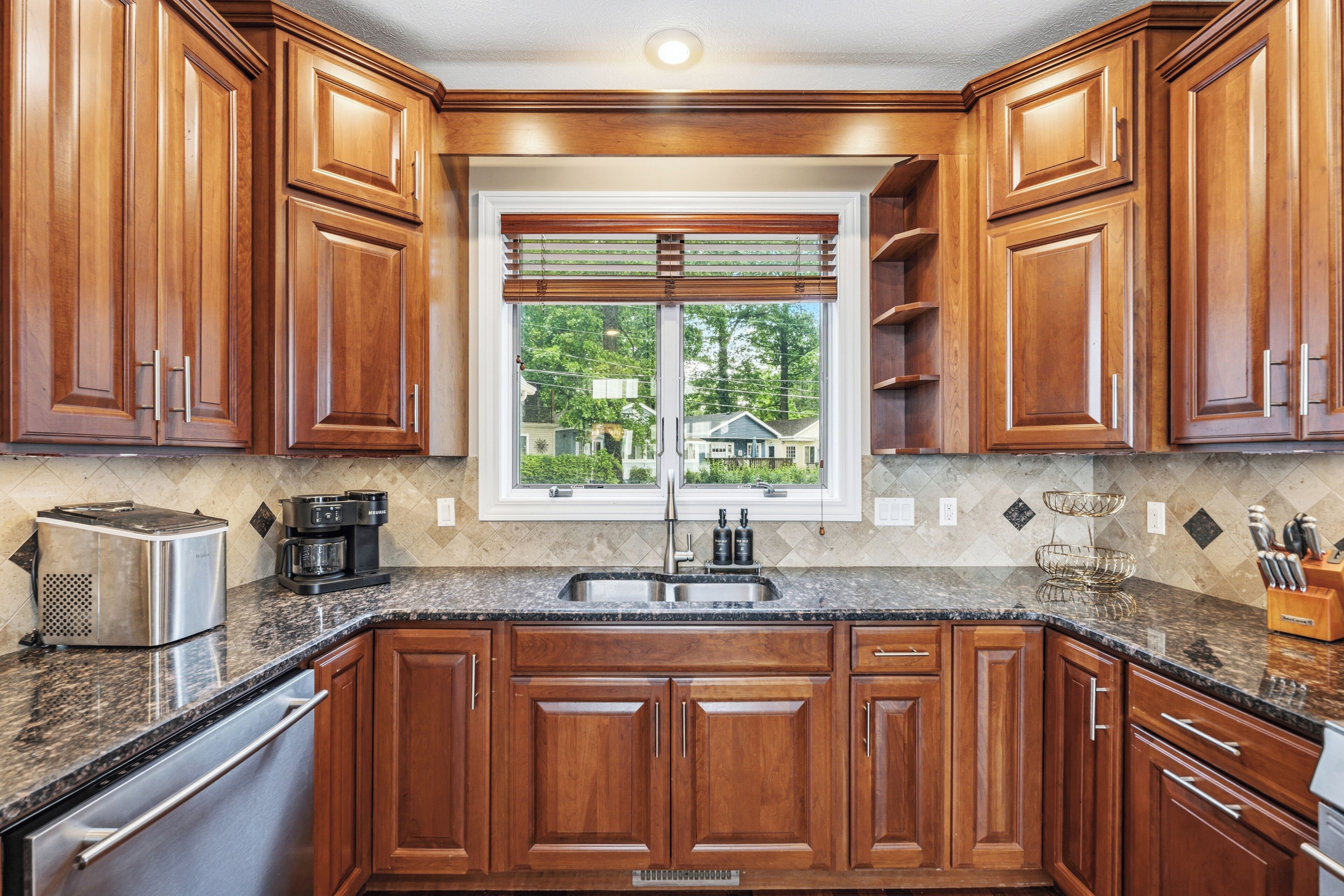 A kitchen with wooden cabinets, a granite countertop, a stainless steel sink, and a window with blinds.