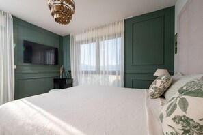 A bedroom with a large bed, a TV mounted on the wall, a window with sheer curtains, and a chandelier. - Villa Olivetum (Okrug Gornji)