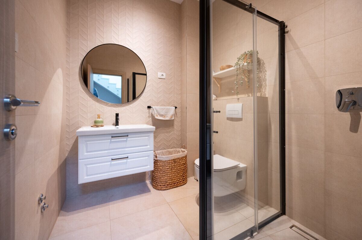 A modern bathroom with a glass shower enclosure, a white vanity with drawers, a round mirror, and a wicker basket.