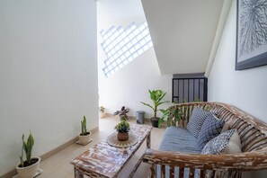 A cozy room with a sloped ceiling, a wicker sofa with patterned cushions, a small round table with a potted plant, and several potted plants on the floor. - Casa Mia Balangan by Betterplace (Jimbaran)