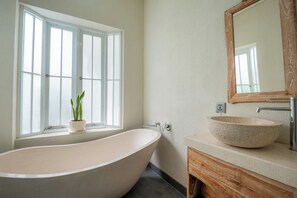 A bathroom with a freestanding bathtub, a wooden vanity, a large mirror, and a window with a potted plant. - Casa Mia Balangan by Betterplace (Jimbaran)