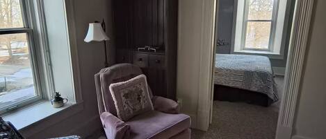 A bedroom with a sloped ceiling, a chair with a cushion, a cabinet, a bed, and a window.