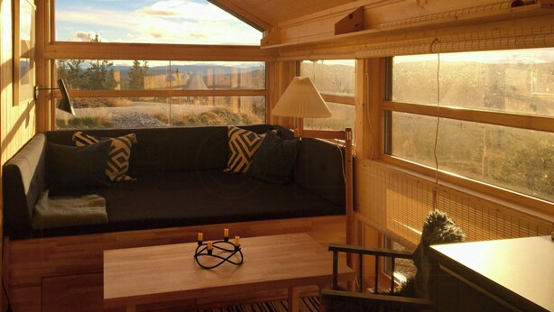 Ein gemütliches Cabin-Interieur mit Holzbalkendecke, einer Couch mit gemusterten Kissen, einem Tisch mit Kerzenhalter und großen Fenstern mit Blick ins Freie.