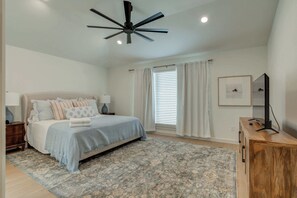 A bedroom with a bed, a ceiling fan, a window with curtains, a bedside table, a lamp, and a TV on a wooden cabinet. - The Foundry - 5BR/3.5BA - Family Retreat (Lubbock)
