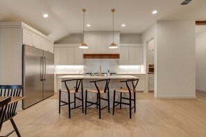 A modern kitchen with white cabinetry, a stainless steel refrigerator, a central island with bar stools, pendant lighting, and a wooden backsplash. - The Foundry - 5BR/3.5BA - Family Retreat (Lubbock)