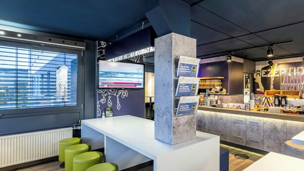 A modern hotel reception area with a large digital screen, a counter with a menu, and green bar stools. - B B Hotel Bremen Sud (Bremen)