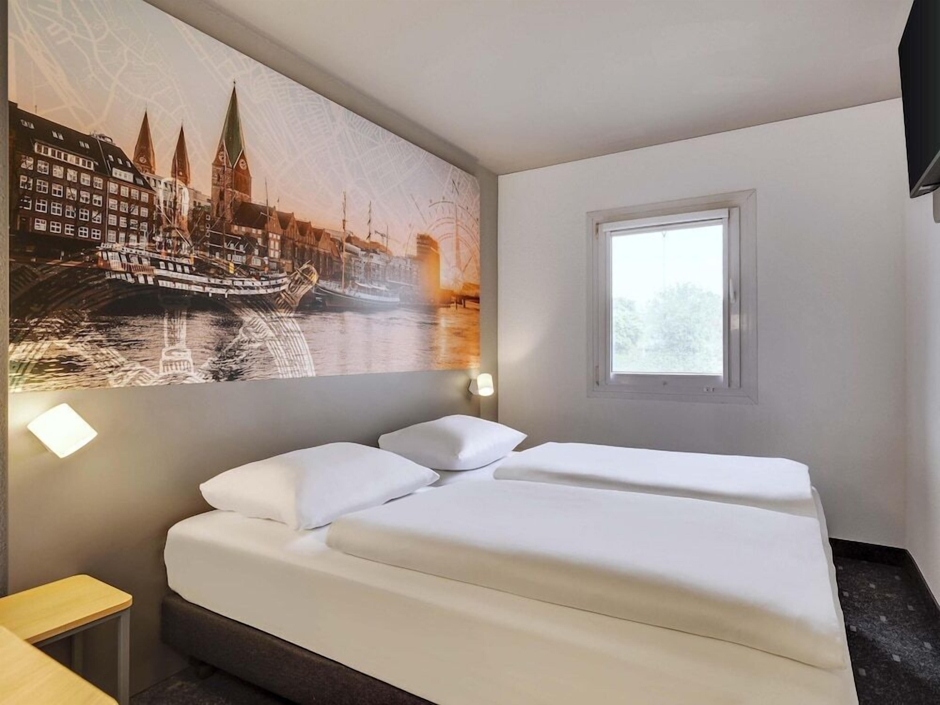 A hotel room with a bed, a window, and a wall with a cityscape painting.