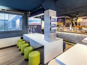 A modern hotel lobby with a reception desk, green bar stools, a large TV screen, and a counter with amenities.