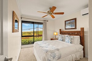 A bedroom with a large bed, a ceiling fan, a nightstand with a lamp, a framed picture on the wall, and a window with a view of greenery. - Ka'anapali Plantation #41 - Vacation West Maui (Lahaina)