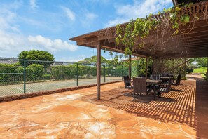 Un patio techado con cancha de tenis, mobiliario exterior y un pérgola cubierta de hiedra.